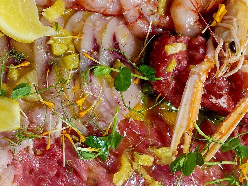 Overhead close-up of a crudo platter with red prawns, langoustines, fish carpaccio and lemon