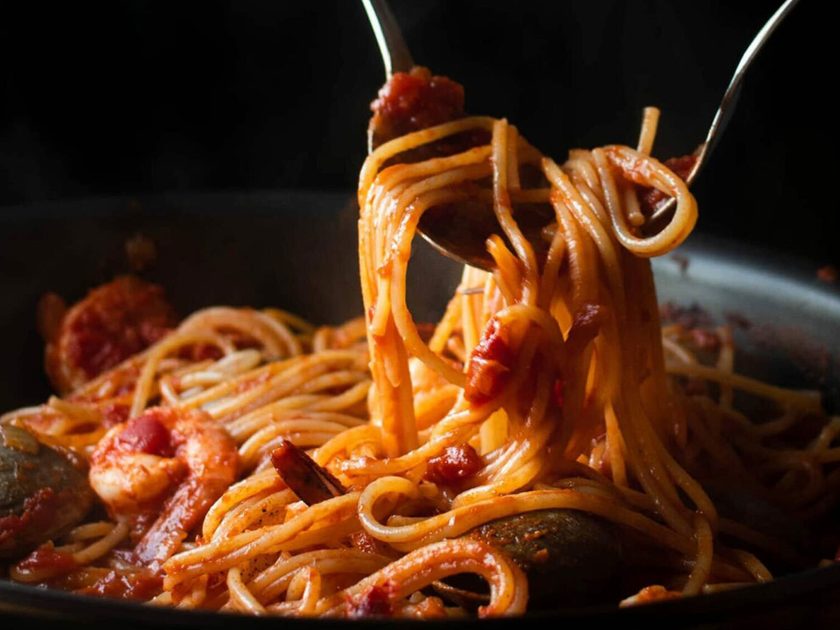 Seafood spaghetti with prawns and clams being lifted from a dark pan in tomato sauce