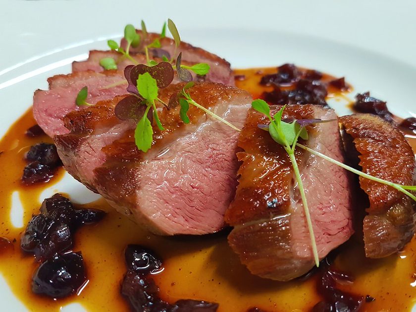 Sliced pan-seared duck breast with dark fruit sauce and microgreens on a white plate