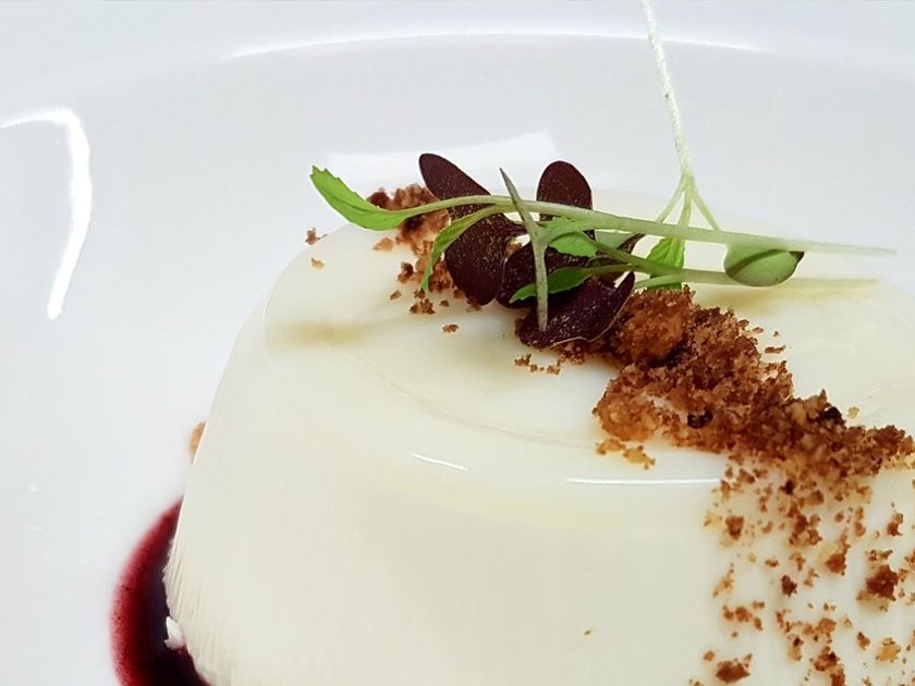 White panna cotta style dessert with dark berry coulis, biscuit crumbs and microgreens on a white plate