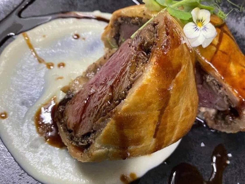 Sliced Beef Wellington on white puree with dark jus and an edible flower on a dark plate